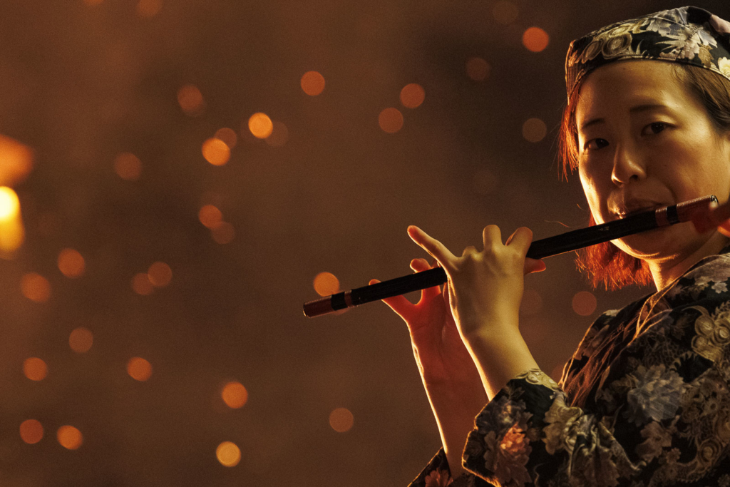 A woman plays a flute with burning embers on the background